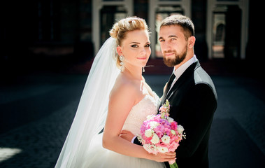 Bearded groom holds bride's waist tender while they pose on the pavement backyard