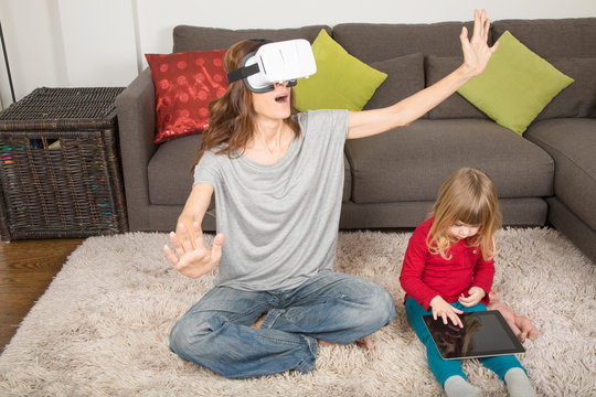 woman with virtual reality headset, or 360 glasses, astonished and three years old child using and watching digital tablet, sitting on carpet indoor home 
