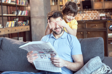 boy closing father's eyes while reading newspaper at home