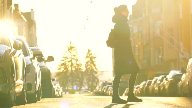 young travel woman in long black coat crossing paved road in the center of town with parked cars on both sides, road, trees and buildings are shining gold under the blind sunlight