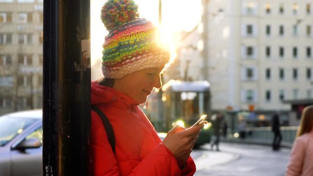 young smiling woman in bright jacket chat using mobile phone while staying on bus station near pole and waiting for friend with urban passing transport on background and under shining bright sunlight