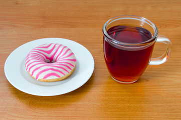Donut with wihte and pink glaze on plate and cup of tea on wooden background.