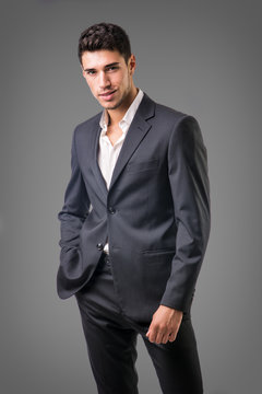 Young businessman confidently posing in front of camera, wearing business suit without neck-tie, with shirt open on neck, on dark background
