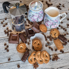 A coffee pot, a sugar bowl, a milk pot, some pieces of chocolate, chocolate sweets, shortbread cookies, coffee beans and star anise on a white wooden background