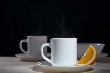 White mug with the steam from the tea next to a slice of orange on a black background