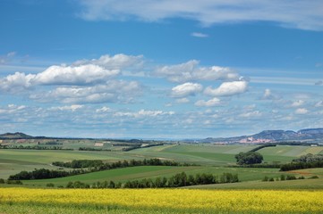 Rural landscape with a cozy village among the hills, fields and meadows under cloudy blue sky
