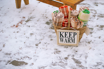 Wicker basket with red, brown and green plaid in a cage in the snow near a wooden bench, and with the words "keep warm"