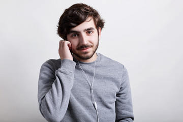 Young handsome bearded hipster with trendy hairstyle standing over white background wearing casual grey sweater enjoying listening to music holding his hand on earphones