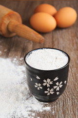 Flour in ceramic bowl, eggs and rolling pin on wooden background