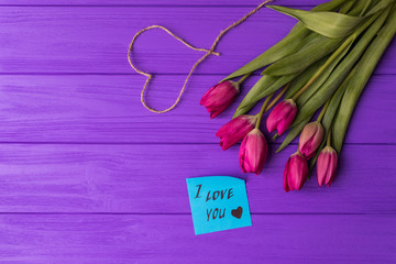Pink colorful tulips over a purple background, in a flat lay composition with heart and inscription on paper i love you.