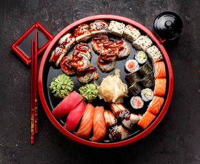 Japanese cuisine. Sushi roll and nigiri set on a round wooden plate with soy sauce and red chopsticks over black concrete background.