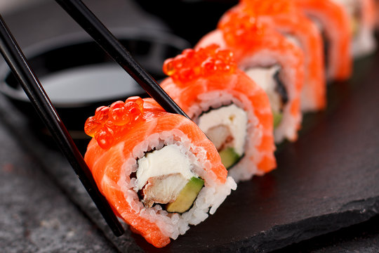 Japanese cuisine. Salmon sushi roll in chopsticks on a stone plate over concrete background.