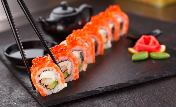 Japanese cuisine. Salmon sushi roll in chopsticks on a stone plate over concrete background.