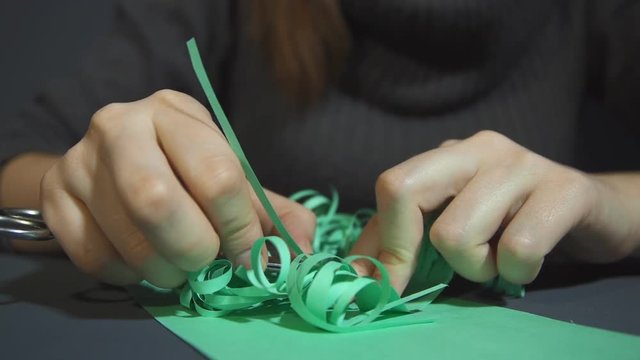 Graceful female hands gently frizzing thinly fringed green paper with metal scissors over a gray background closeup