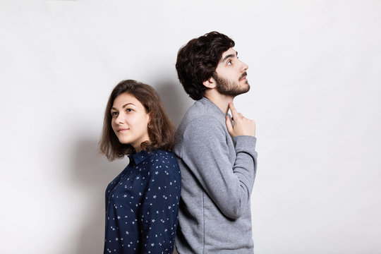 A Horizontal Portrait Of Two Young People Standing Back To Back Having Thoughtful Expression. A Stylish Young Bearded Man Holding His Finger On His Neck Looking Up Thinking Standing Back To His Girl