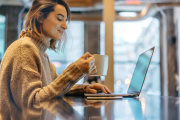 Young smiling beautiful woman drinking coffee, Woman wearing in cozy sweater and using modern laptop, blurred background, shallow DOF