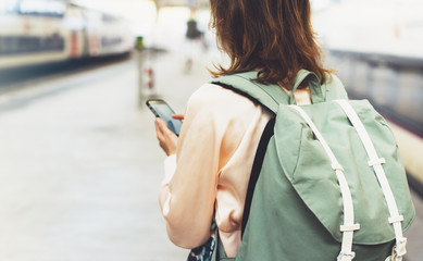 Obraz premium Enjoying travel. Young pretty woman waiting on the station platform with backpack on background electric train. Tourist texting message and plan route of railway, railroad transport concept