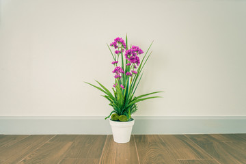 artificial plant with purple flower on wooden floor and white cement wall textured background