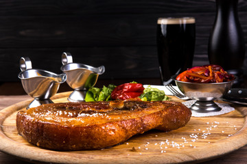 Roasted slice of pork leg with bone. Two sauces, Red beans, onion, bell pepper on wooden background and black glass of beer. Food cocept.