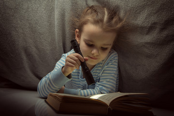 Little girl reading fairy tales book under the covers at the evening with lantern. Cute kid playing before going to sleep, image toned.