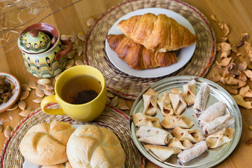 Black tea in yellow mug with croissants, buns and homemade cookies on wooden table