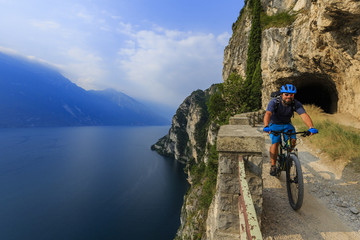Fototapeta premium Mountain biking at sunrise man over Lake Garda on path Sentiero della Ponale, Riva del Garda, Italy
