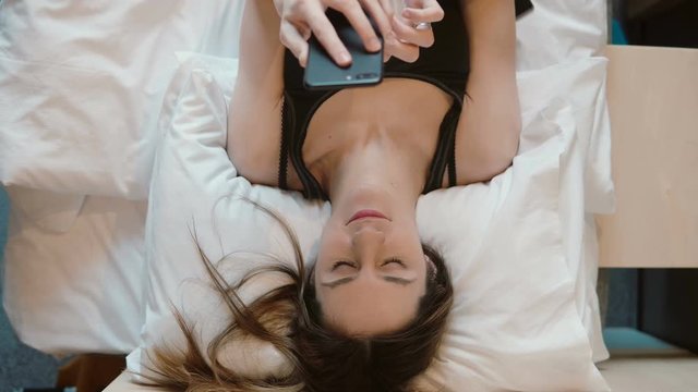 Young woman lying on the bed and holding smarphone. Girl browsing the Internet, chatting with someone.
