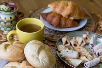 Black tea in yellow mug with croissants, buns and homemade cookies on wooden table