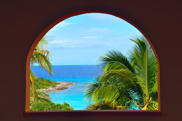 view of tropical beach and palm trees through the window in the form of an arch. Beautiful sunny day, emerald water, nobody.
