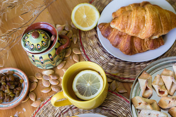 Black tea with lemon in yellow mug with croissants, buns and homemade cookies on wooden table