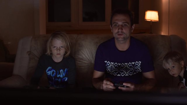 Middle-aged father in blue tshirt playing console with his blonde children sitting at sofa in the evening