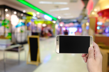 Right hand using smart phone with blank screen on abstract blur background of storefront in fashion mall or department store.