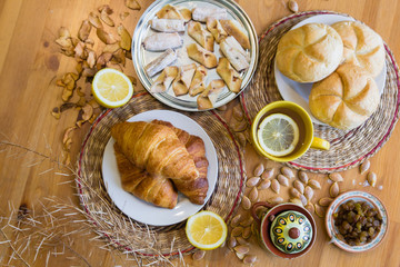 Black tea with lemon in yellow mug with croissants, buns and homemade cookies on wooden table
