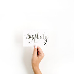 Fototapeta premium Minimal pale composition with girl's hand holding card with word Simplicity written in calligraphic style on paper on white background. Flat lay, top view