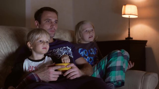 Middle-aged male in blue t-shirt sitting at sofa with two blonde sons watching TV eating candies from bowl