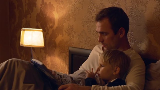 Middle-aged father in white t-shirt reading book of fairy tales to his blonde child lying in bed close to torchere in slowmotion