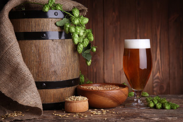 Old barrel, glass of dark beer and its ingredients on table against blurred wooden background