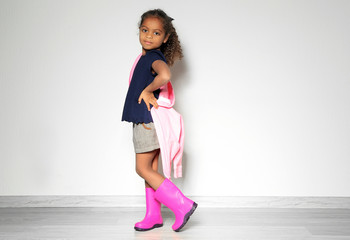 Cute little African American girl in rubber boots against light wall. Fashion concept