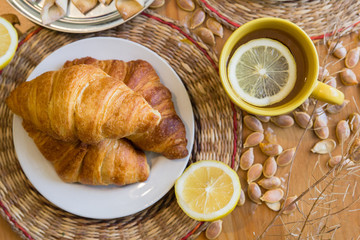 Black tea with lemon in yellow mug with croissants, buns and homemade cookies on wooden table