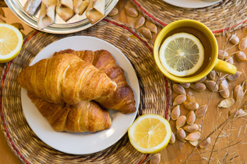 Black tea with lemon in yellow mug with croissants, buns and homemade cookies on wooden table