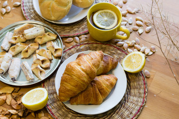 Black tea with lemon in yellow mug with croissants, buns and homemade cookies on wooden table