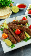 Homemade Kofta kebabs on skewers with pita, lime, vegetables, sweet chili and yogurt sauce on white plate.
