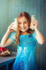 Сhild girl in blue dress listens to a sea shell and laughs.