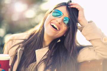 Portrait of a young woman with sunglasses, laughing outside, holding coffee to go