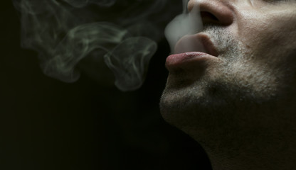  Close up shot of a man exhaling cigarette smoke on black background with dramatic light.