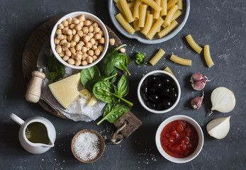 Ingredients for cooking rigatoni pasta with chickpeas, spinach and olives in a tomato sauce on a dark background, top view. Vegetarian food concept