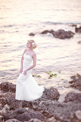 Fototapeta na wymiar Young Blond Bride in White Dress Posing by the Sea on Sunset
