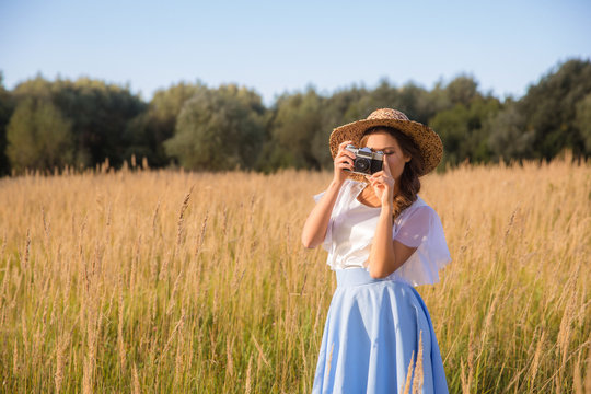 Girl in hat shooting photo walking in golden dried grass field with camera