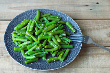 Green beans mixed with sesame seeds served on a gray plate with fork on wooden table. Diet eating concept