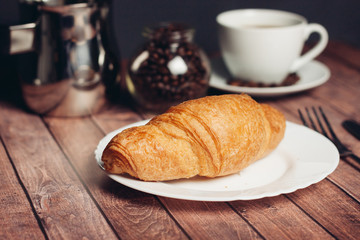 Croissant in a white plate, a jar with coffee beans, a cup in the background